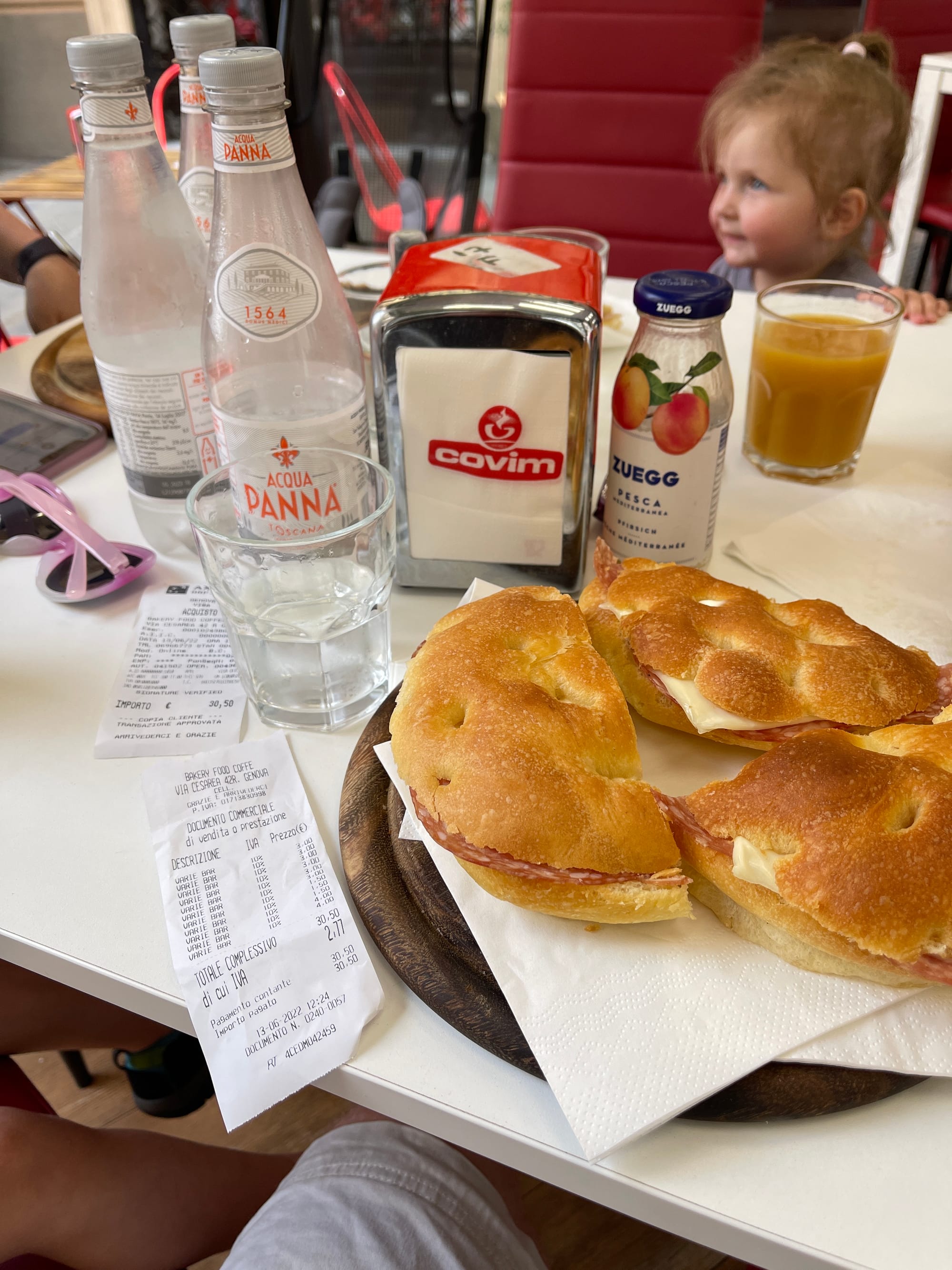 Fresh focaccia sandwiches with meat and cheese on plate at Italian cafe table with Acqua Panna water bottles and receipt, young child in background