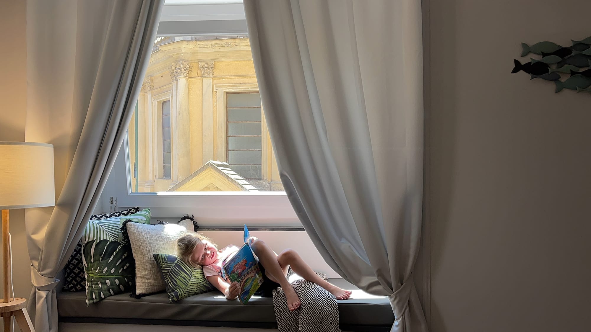 Young girl reading book while sitting in bay window seat overlooking historic buildings in Genoa, Italy